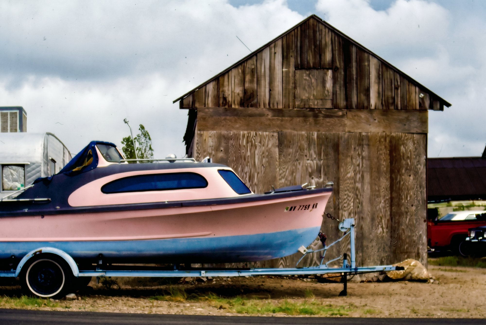Pink-and-blue vintage boat on a trailer beside a weathered wood shed under a cloudy Nevada sky.